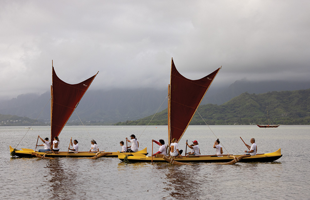 wide shot of two boats with red sails and a line of people rowing